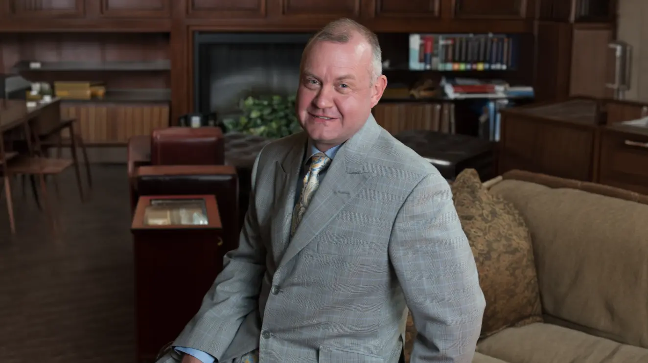 Jeff Snell, IBBA Hall of Fame business intermediary and ENLIGN leader, smiling in a gray suit seated in a wood-paneled office with a fireplace and bookshelves.