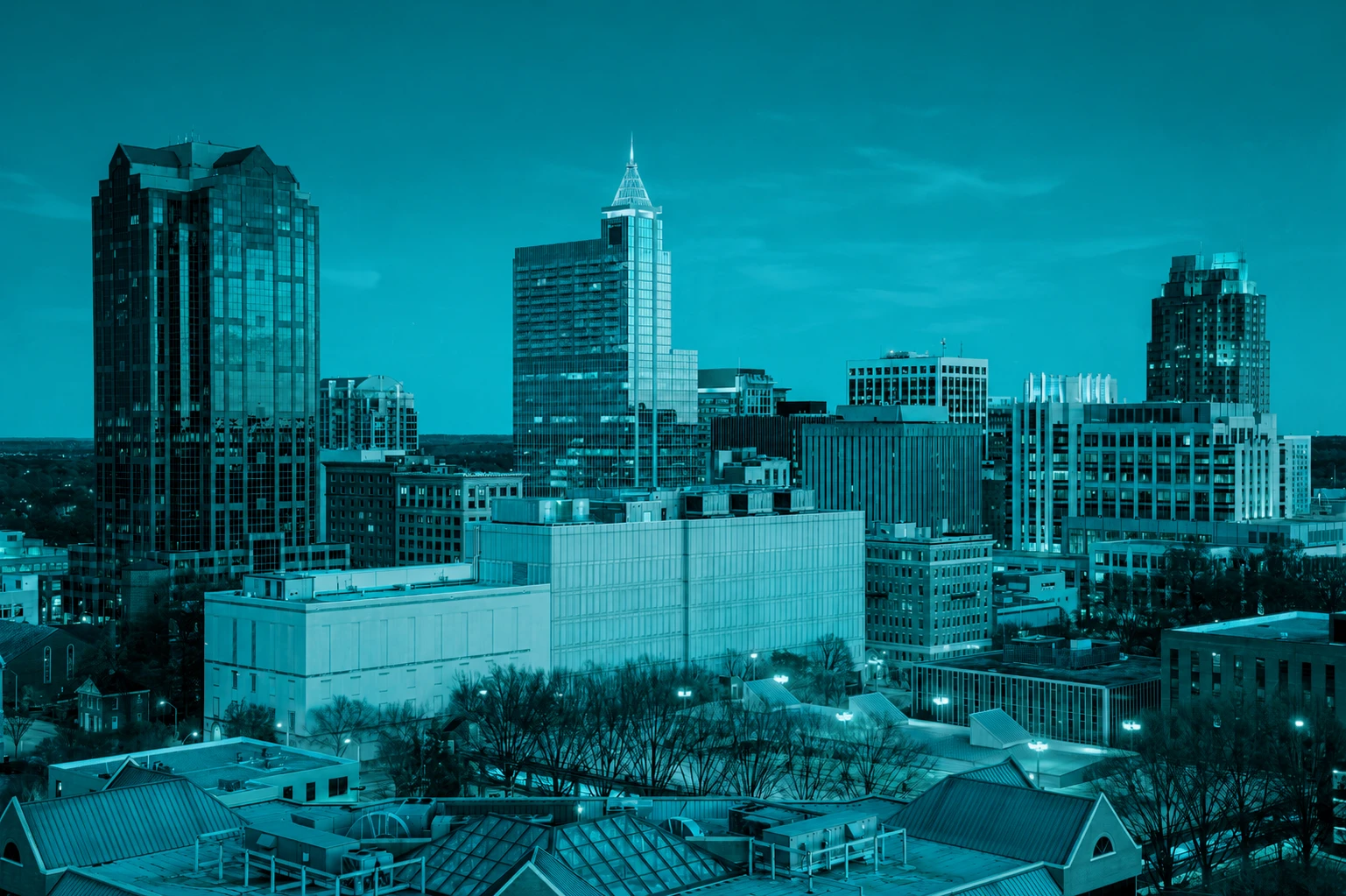 A monochromatic teal-toned cityscape showing modern high-rise office buildings and skyscrapers of a downtown business district against an evening sky.