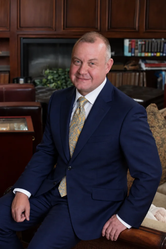 Jeff Snell, IBBA Hall of Fame business intermediary and ENLIGN leader, smiling in a blue suit, white shirt and gold paisley tie, seated in a wood-paneled office with a fireplace and bookshelves.