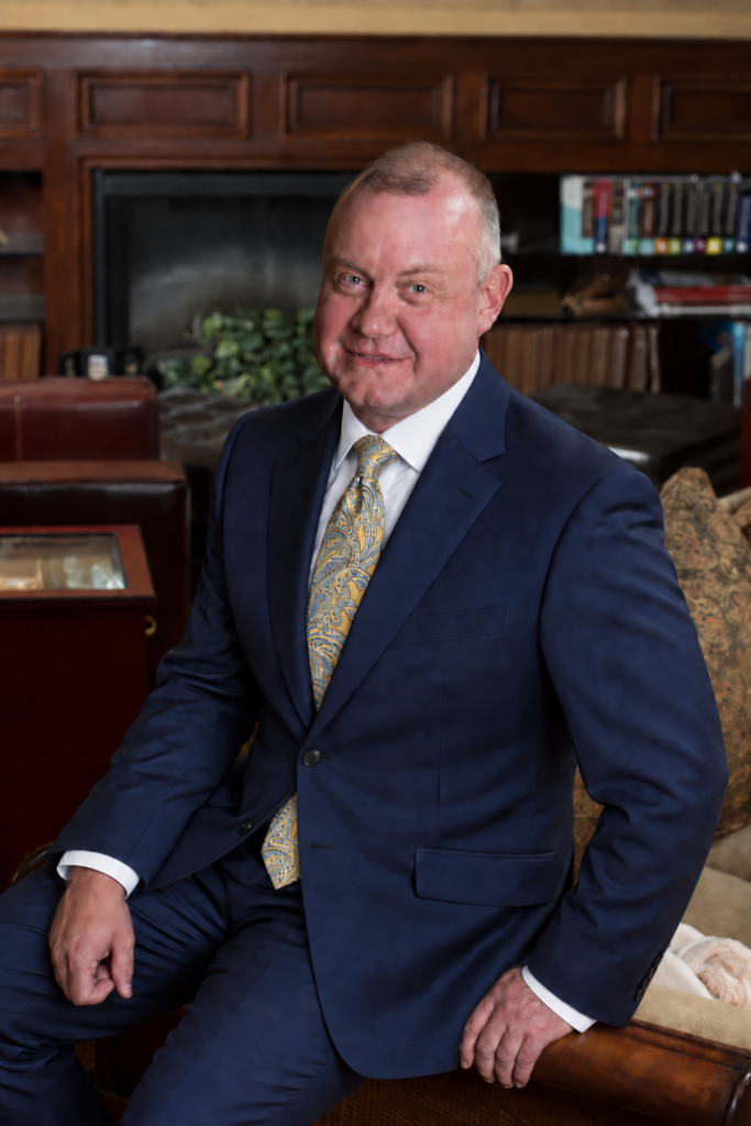 Jeff Snell, IBBA Hall of Fame business intermediary and ENLIGN leader, smiling in a blue suit, white shirt and gold paisley tie, seated in a wood-paneled office with a fireplace and bookshelves.